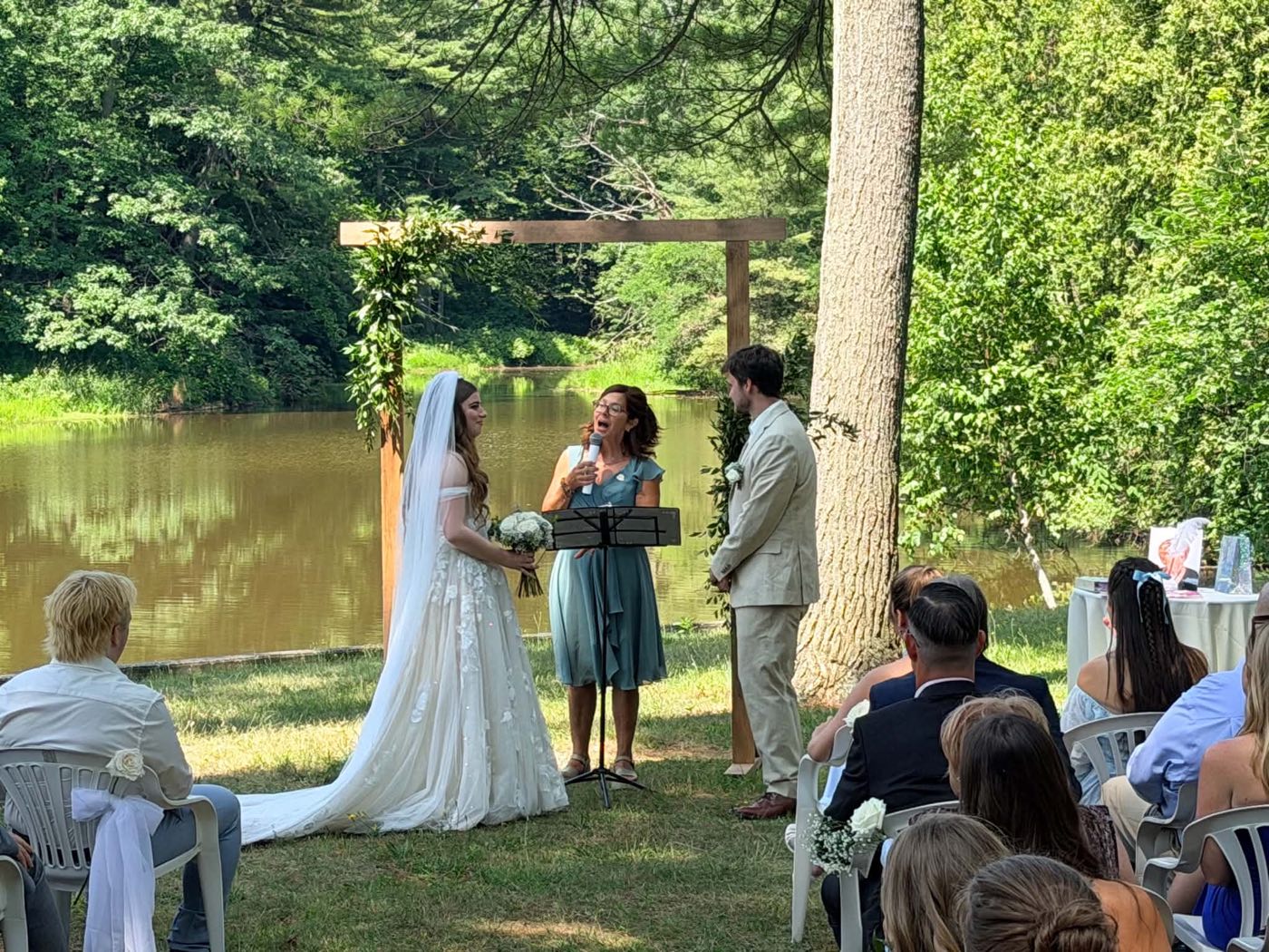 Cérémonie de mariage au bord d’un étang, Isabelle Ferland au micro sous une arche de verdure.