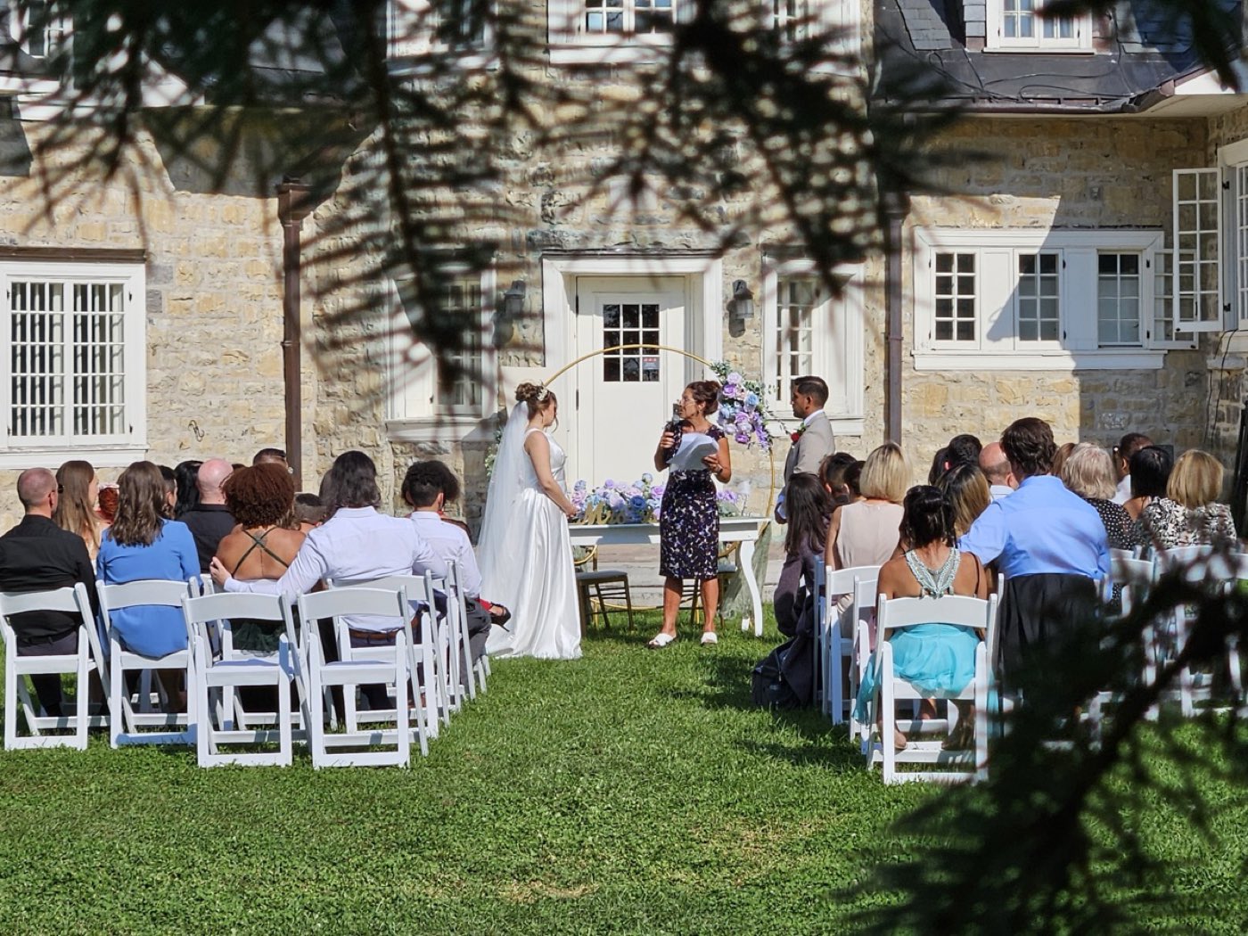 Cérémonie de mariage devant un manoir de pierre, les invités assis sur la pelouse.