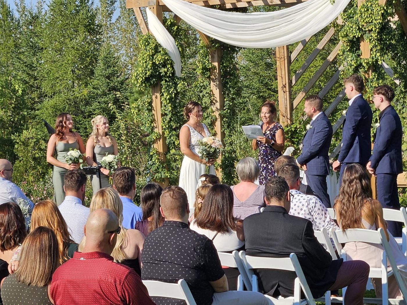 Cérémonie de mariage extérieure sous une pergola couverte de houblon et de voiles blancs.